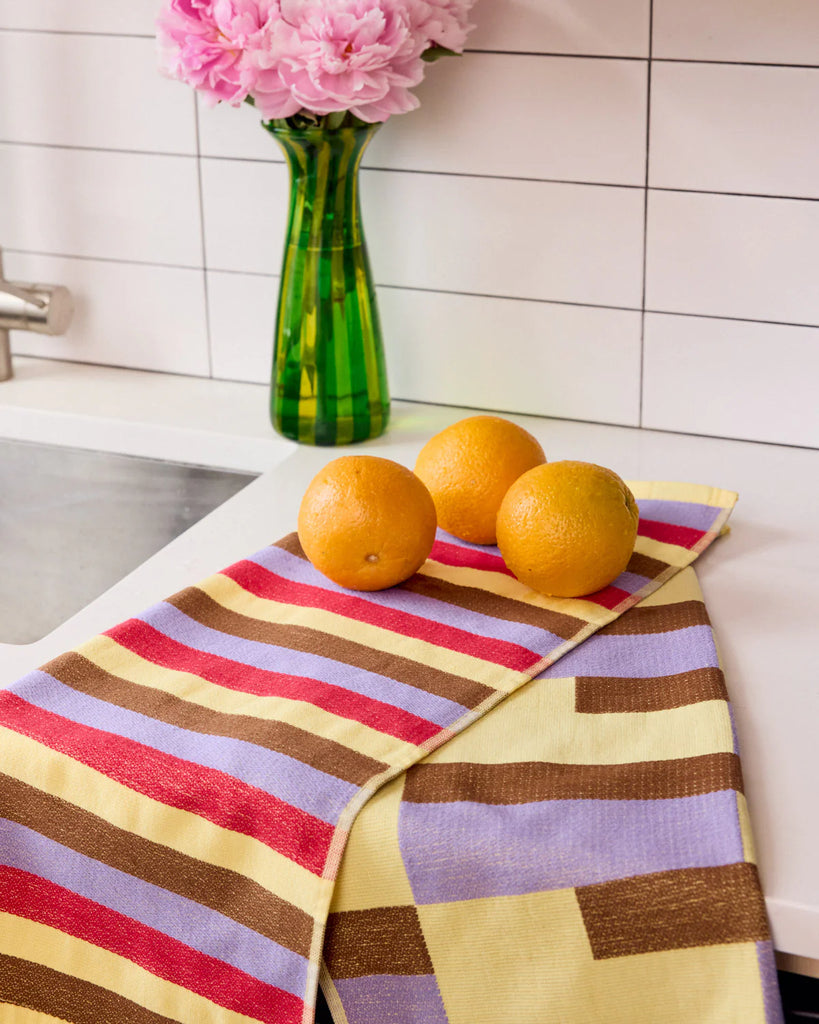 Two pink,lilac,brown and yellow striped towels with oranges and a vase of pink flowers on a kitchen counter.
