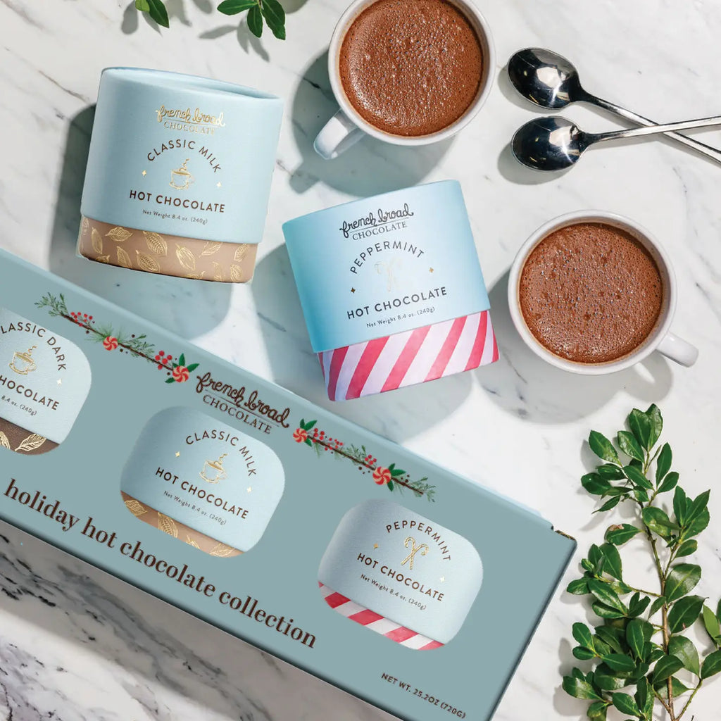 Hot chocolate packaging with mugs and spoons on a marble surface.  Classic Milk, Dark & Peppermint Hot Chocolate in three 8.4oz cylinders.