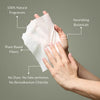 A person holds a translucent white facial mask sheet against a muted gray-green background, with callout text reading '100% Natural Fragrances,' 'Nourishing Botanicals,' 'Plant-Based Fibers,' and 'No Dyes. No fake perfumes.