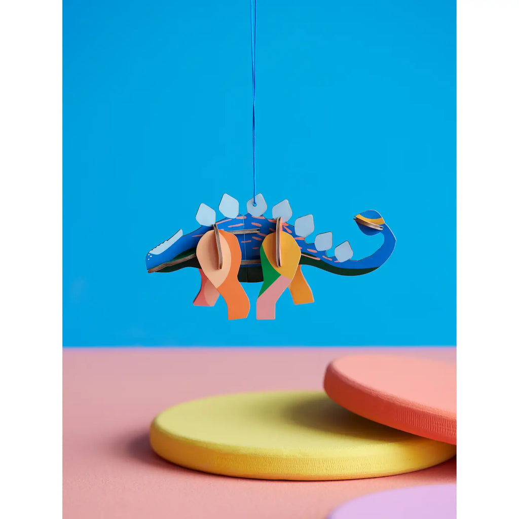Colorful paper dinosaur hanging against a blue background with pink and yellow platforms at the bottom.