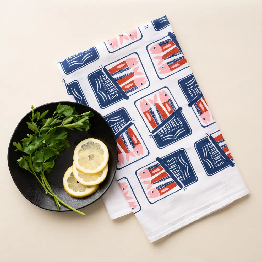 Tea towel with tinned fish design next to a plate with lemons and herbs on a beige background